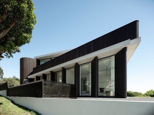 A contemporary multi-level residential facade with strong horizontal cantilevered concrete slabs, dark vertical ribbed cladding, tall floor-to-ceiling glazing, and curved end walls. The house sits behind a low pale rendered boundary wall and dark metal fencing, with landscaped greenery surrounding it.