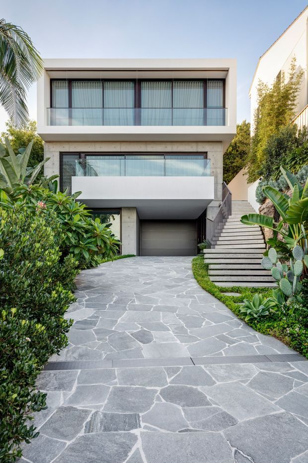 Front exterior view of a contemporary multi-storey residence with a clean rectilinear facade, broad glazed balcony, white rendered walls, and a projecting upper volume. A grey stone-paved driveway leads to an integrated garage, with a wide exterior stairway rising alongside the house. Lush tropical and subtropical planting frames the entry.