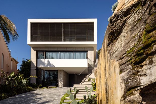 Contemporary multi-storey residence viewed from the driveway, tightly positioned beside a dramatic natural rock escarpment. The facade combines a crisp white rectangular upper volume, broad dark louvred glazing, a recessed glass-fronted middle level, and a projecting balcony. Stone paving, stepped landscaping, and cactus planting lead to the entrance and garage-like lower opening. Bright sunlight and a clear blue sky emphasize the modern geometric form.