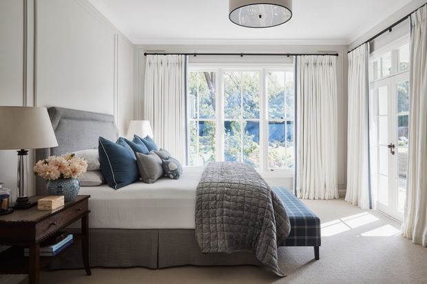 A bright, refined bedroom featuring a large upholstered bed with layered blue and grey cushions, a quilted grey throw, and a blue plaid bench at the foot. Tall white-framed windows and glazed French doors with full white curtains bring in abundant natural light. The room includes a dark timber bedside table, table lamp, floral arrangement, and a simple flush-mount ceiling light. The overall look is classic and calm with soft neutral finishes and blue accents.