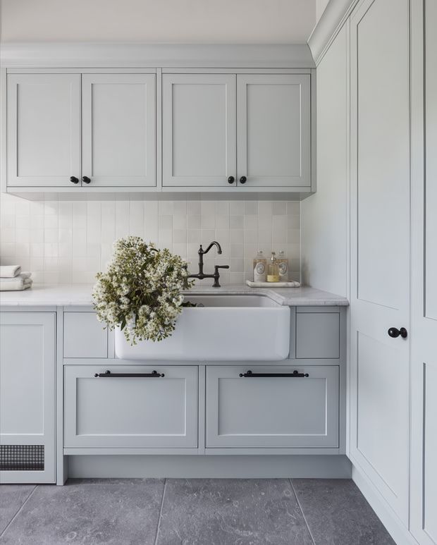 A refined laundry room with pale grey Shaker-style cabinetry, integrated overhead cupboards, a deep white farmhouse sink, dark traditional tapware and handles, white square tiled splashback, and large grey stone-look floor tiles. A tall bank of matching storage cupboards lines the right wall. Soft natural light and restrained styling create a calm, classic country-inspired interior.
