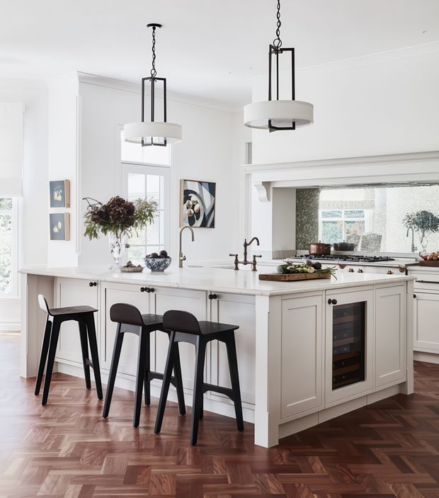 Bright, refined kitchen interior with a large white island, integrated wine fridge, paneled cabinetry, dark bar stools, and twin pendant lights. The room features white walls and joinery, dark herringbone timber flooring, a rear cooking area, and classic architectural trim around the opening. Natural light enters through tall windows, creating an elegant, welcoming atmosphere.