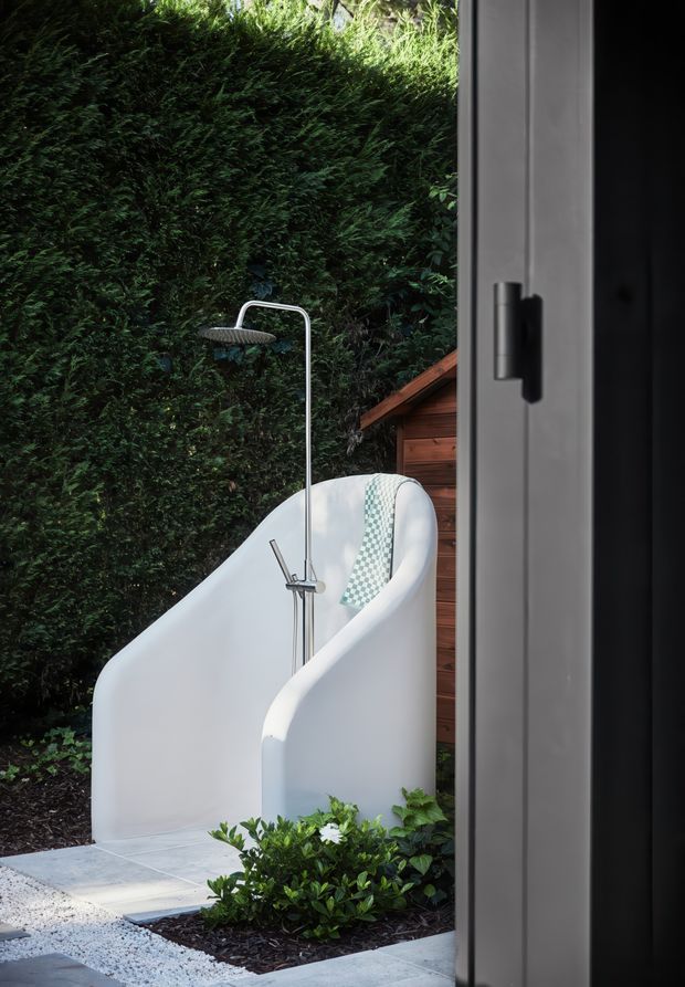 An outdoor bathroom-style setting viewed from a doorway, featuring a freestanding white sculptural bathtub with an integrated shower fixture. The tub sits on pale tiled paving beside dark mulch and clipped greenery, with a dense evergreen hedge forming the backdrop and a warm timber-clad structure behind it. The image has a refined, private, nature-integrated feel with strong contrast between white sanitaryware, green planting, and timber.