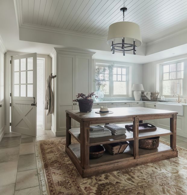 A bright, refined kitchen with pale built-in cabinetry, a large rustic timber island with open shelving, a patterned area rug, and a large drum pendant light. The room features extensive multi-pane windows, a glazed panel door, and a white beadboard-style ceiling with traditional trim.