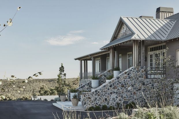 Exterior view of a refined single-storey rural-style house on a gently elevated site, featuring a steep gabled metal roof, covered verandah, vertical timber cladding, pale trim, and substantial irregular natural-stone retaining walls and steps. Sparse landscaping and a broad open landscape surround the home.