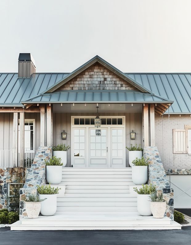 Front exterior view of a symmetrical, light-toned building entrance with a broad gabled porch, standing-seam metal roof, shingle-clad gable, painted timber detailing, glazed double doors, and wide white steps. Stone-clad side walls and large white planters frame the entry.