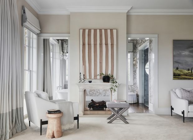 Elegant light-filled living room with tall windows dressed in full-length curtains, a white upholstered armchair and sofa, pale carpet, a decorative fireplace with ornate mantel, and a central upholstered cross-leg ottoman. The room features refined wall mouldings, broad crown cornices, and a neutral taupe, white, and soft grey palette, with abstract artwork and restrained decorative styling.