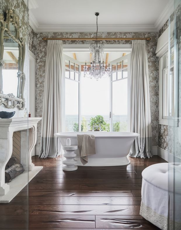 Elegant, richly decorated bathroom with a freestanding white bathtub centered before tall glazed doors overlooking a bright exterior. The room features dark polished timber flooring, ornate grey patterned wallpaper, elaborate white cornices and mouldings, full-height pleated curtains, a crystal chandelier, a decorative fireplace with an oversized ornate mirror, and a partial upholstered seating or bed-like element at right. The overall aesthetic is luxurious, traditional, and refined, with abundant natural light and ocean or landscape views beyond the glazing.