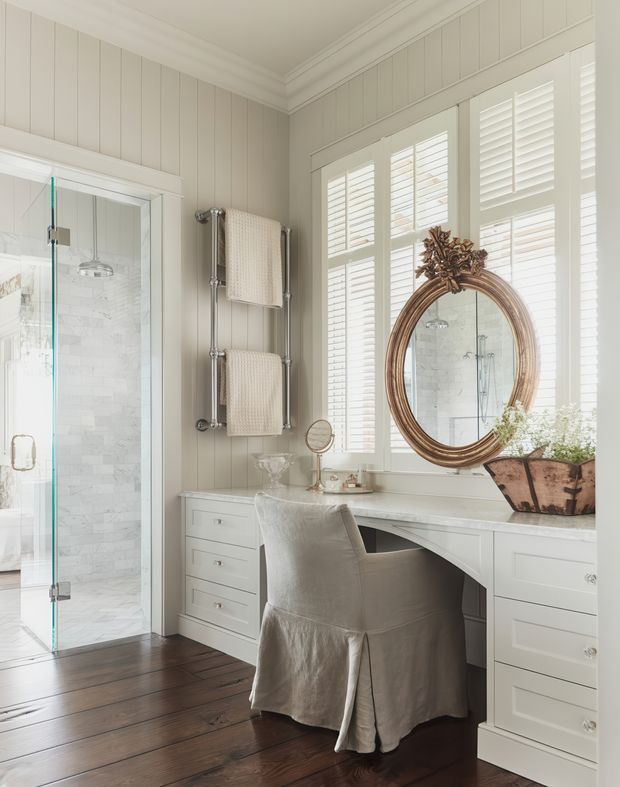 A refined bathroom with a built-in white vanity and dressing desk beneath plantation-shuttered windows. The room features pale vertical wall panelling, detailed crown moulding, a frameless glass shower with white marble-look tile, a polished chrome heated towel rail, dark timber flooring, and an ornate round bronze-framed mirror with floral detailing. A fabric-covered chair and decorative accessories create a softly traditional, elegant atmosphere.