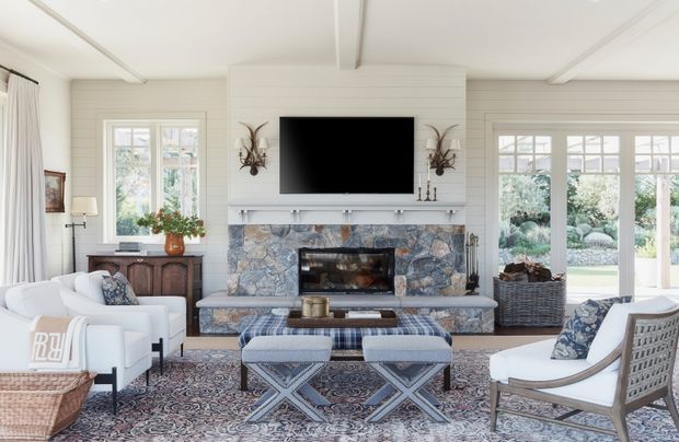 Bright, refined living room with white painted horizontal wall panelling, a substantial natural-stone fireplace surround, and a white mantel supporting a wall-mounted television. The room includes upholstered seating, patterned ottomans, a large patterned rug, a timber sideboard, tall French-style glazed doors, and garden views. The ceiling has simple recessed panel detailing and exposed-looking beams, creating a classic coastal country-house atmosphere.