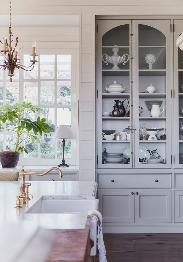 Bright, refined kitchen interior featuring a large white marble island with an undermount sink and brass bridge faucet, pale grey-blue cabinetry, glass-front display cupboards, white horizontal wall panelling, and a large multi-pane window. The cabinetry displays decorative china and silverware, while a traditional brass chandelier and potted plant add warmth. The overall composition is light, classic, and coastal-inspired.
