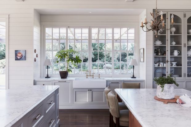 Bright, refined kitchen interior with white horizontal wall panelling, a large multi-pane bay-style window, pale cabinetry, marble-look island and dining bench surfaces, dark timber flooring, and glass-fronted display cabinetry. A chandelier, table lamps, greenery, and upholstered seating add a traditional yet relaxed character.