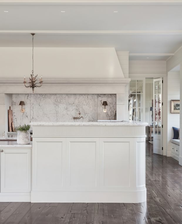 Bright, refined kitchen interior dominated by a large white island with paneled cabinetry and a fluted apron, a veined marble backsplash, pale wall panelling, integrated cabinetry, dark timber flooring, and a traditional chandelier. Glazed French doors open toward an adjoining room, with a glimpse of seating beyond.