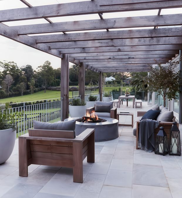 A spacious outdoor terrace beneath a substantial timber pergola, furnished with contemporary lounge seating, a central round fire pit, occasional tables, potted plants, and decorative lanterns. Pale large-format paving contrasts with dark stained timber beams and charcoal furnishings. The terrace overlooks a landscaped garden and appears connected to a glazed building facade.