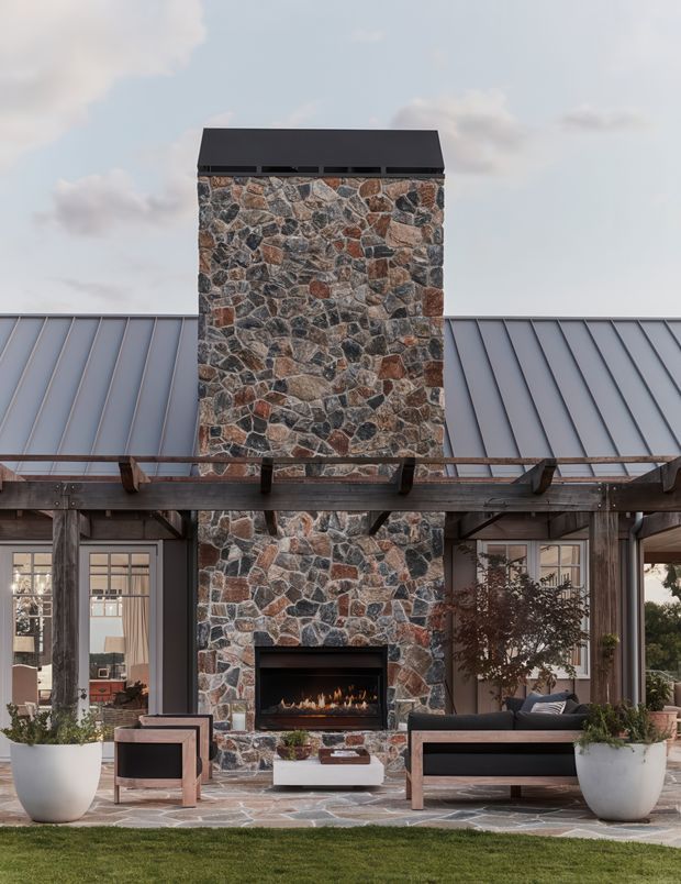 Exterior view of a sophisticated single-storey home centred on a tall, irregular natural-stone chimney and built-in outdoor fireplace. The facade combines dark standing-seam metal roofing, timber-framed glazing, exposed timber pergola structure, and a stone-paved terrace furnished with outdoor sofas and planters. The composition is symmetrical and contemporary-rustic, with a lawn and open sky in the background.