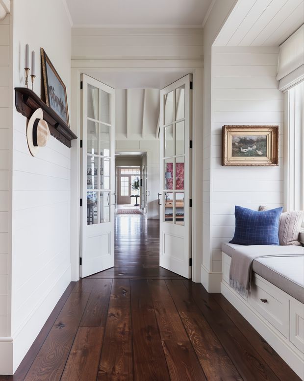 Bright traditional coastal-style hallway or landing with dark wide-plank timber flooring, white horizontal wall panelling, and white French doors opening toward adjoining rooms. A built-in window seat with drawers and cushions occupies the right side, while framed artwork and a decorative hat shelf line the left wall. The view extends through multiple glazed doorways toward a light-filled interior with exposed ceiling beams.