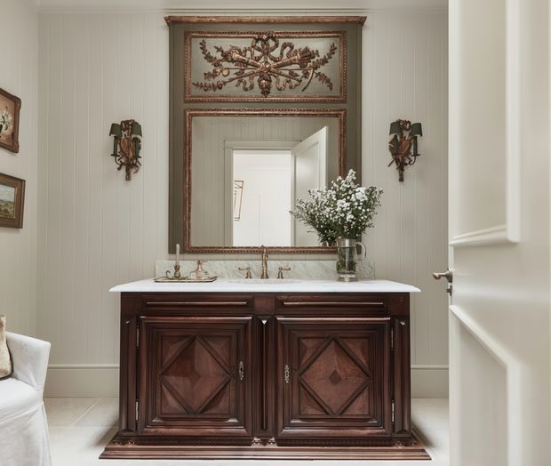 Elegant traditional bathroom vanity area with a dark stained, heavily carved timber cabinet, pale stone benchtop and integrated basins. The room has vertical wall panelling, light tiled flooring, ornate framed mirror and matching antique-style wall sconces. A partially open white door and a glimpse of an upholstered chair are visible at the edges.