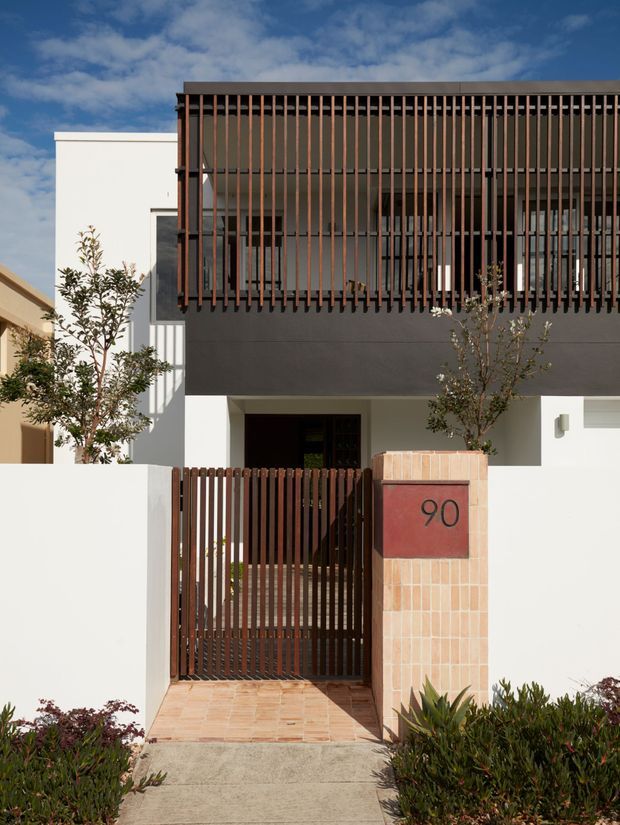 Front exterior view of a contemporary multi-storey residence with a white rendered facade, dark charcoal upper wall, and prominent vertical timber-toned screening over the balcony and windows. A matching slatted pedestrian gate leads to the recessed entry, beside a tiled address pillar marked 90. The property includes a paved walkway and restrained landscaped planting under a partly cloudy sky.