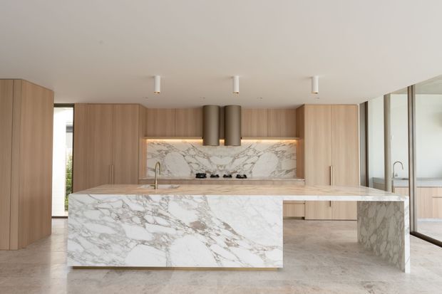 A spacious contemporary kitchen featuring a large waterfall island clad in heavily veined white and grey natural stone, matching stone splashback, and light timber cabinetry. A dark cylindrical rangehood sits above the cooktop, with integrated appliances and minimal linear lighting. Full-height glazing or reflective panels line the right side, creating a bright, refined interior.