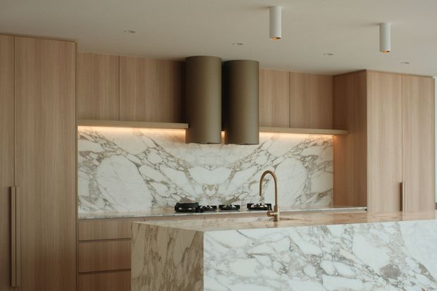 Contemporary kitchen featuring full-height light timber cabinetry, a dramatic white marble splashback and waterfall island, integrated warm under-cabinet lighting, a gas cooktop, and twin cylindrical bronze-toned rangehood elements. The palette is warm and refined with clean, minimal detailing.