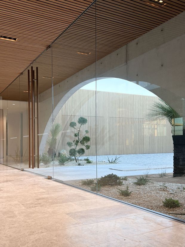 A refined contemporary interior corridor or foyer with expansive frameless glass walls overlooking a minimalist landscaped courtyard. The space features a warm slatted timber ceiling, pale large-format stone or porcelain floor tiles, a sculptural curved concrete arch opening, and slender vertical timber elements near the glazed entry. The courtyard includes pale gravel, sculpted planting, and a ribbed light-coloured architectural wall, creating a calm indoor-outdoor connection.
