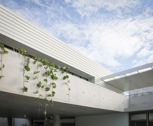 Modern minimalist residential exterior courtyard or terrace view, featuring smooth pale concrete walls, a projecting cantilevered upper volume, horizontal light cladding, slim dark-framed glazing, and a narrow open pergola-like roof structure. Hanging trailing greenery softens the architectural concrete surfaces beneath a bright partly cloudy sky.