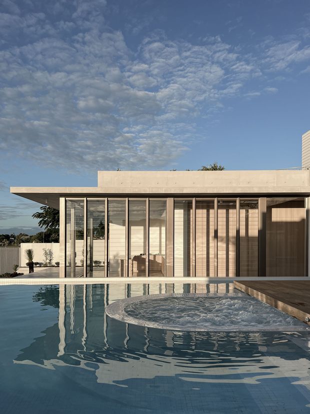Exterior architectural photograph of a sleek modern residence beside a swimming pool. The low, horizontal building has a broad flat roof, extensive floor-to-ceiling glazing with dark slender frames, pale rendered walls, and timber-toned vertical screening. The foreground pool includes a circular spa section and strongly reflects the facade and sky.