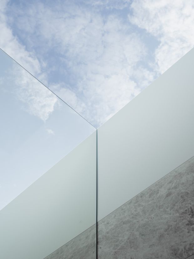 Abstract architectural detail showing two pale, smooth wall planes meeting at a sharp vertical corner, with a reflective glass or glazed plane on the left. A grey textured stone or concrete surface rises diagonally from the lower right, while an open sky is visible above, creating a minimalist geometric composition.