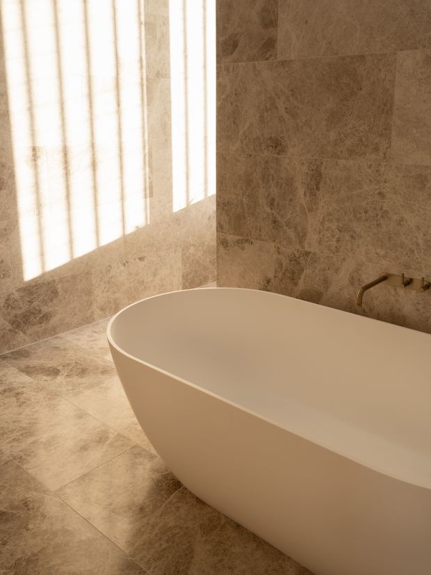 A warm, softly lit bathroom featuring a freestanding sculptural white bathtub against walls and flooring clad in large-format beige-brown stone-look tiles. A wall-mounted brass-toned bath filler is visible, with strong sunlight filtering through a window and casting vertical shadows across the tiled surfaces.