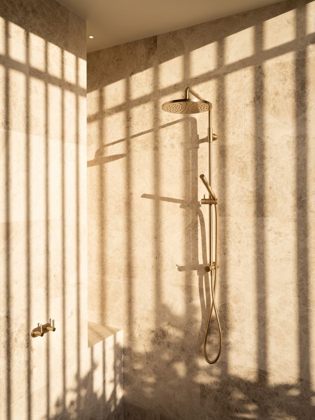 A minimalist walk-in shower enclosed by warm beige, stone-look wall and floor finishes. A brushed brass rainfall showerhead, handheld shower, controls, and exposed plumbing are mounted against the tiled wall. Strong sunlight casts geometric window-frame shadows across the surfaces, creating a warm, calm atmosphere.