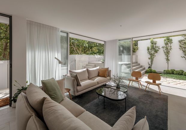 Bright contemporary living room with large corner glazing opening to a landscaped courtyard. Two neutral upholstered sofas surround a black oval glass coffee table, with a pair of timber lounge chairs near the windows. Sheer curtains, pale tiled flooring, a dark area rug, and white walls create a calm, airy interior; concrete steps and planted courtyard walls are visible outside.