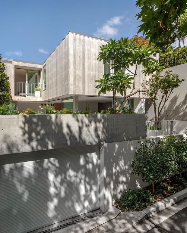 A contemporary multi-level residential building viewed from the street, featuring a minimalist rectilinear form, a pale vertically textured concrete or rendered facade, recessed glazing, balconies with slender metal balustrades, and an integrated garage at street level. Mature tropical-style planting and a raised landscaped terrace soften the clean architectural lines.