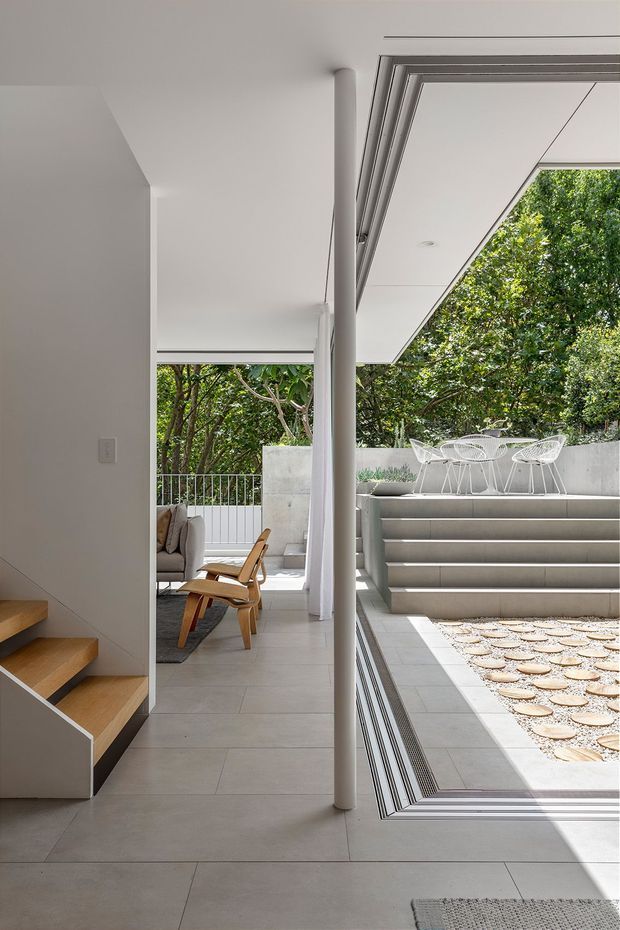 A refined contemporary interior opening onto a landscaped sunken courtyard. The image shows a pale tiled hallway or living area with a sculptural white column, timber stair treads, a partially visible lounge chair and sofa, and extensive frameless or slim-framed glazing. Outside, broad concrete steps lead to a raised patio furnished with white wire-frame dining chairs and a table, surrounded by mature greenery. The architecture is clean-lined and minimalist, with strong indoor-outdoor connection and abundant natural light.