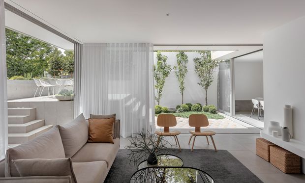 Bright contemporary living room opening onto a landscaped courtyard, with extensive floor-to-ceiling glazing, sheer curtains, a neutral sofa, timber lounge chairs, a dark rug, and a round glass coffee table. A raised outdoor terrace and steps are visible to the left, while the courtyard garden and additional seating are visible through the open glazing.