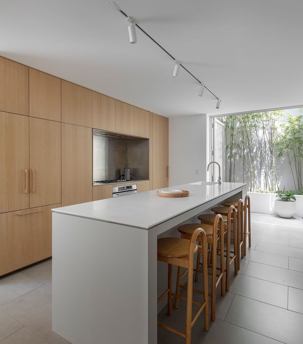 Bright contemporary kitchen with a long white island and integrated seating for four or more, light timber full-height cabinetry, a recessed cooking niche, stainless-steel tapware, pale tiled flooring, and track lighting. Glazed doors open toward a planted courtyard with bamboo, bringing abundant natural light into the space.