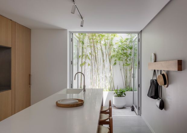 A compact, refined kitchen viewed toward full-height glazed doors opening to a bamboo-filled courtyard. The room features a long pale stone island with an integrated sink and curved metal tap, light timber cabinetry with integrated appliances, grey tiled flooring, white walls, and a simple wall-mounted timber shelf with hooks. Track lighting runs across the ceiling, and stools line the island.