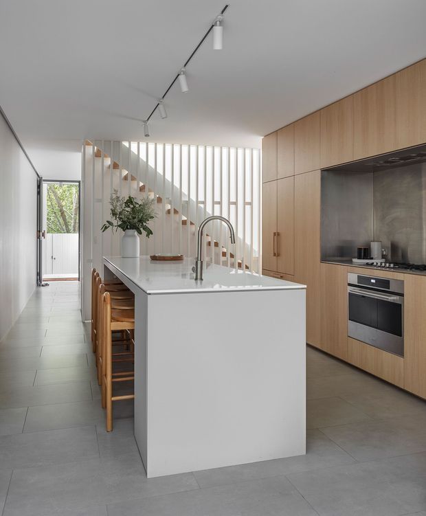 Contemporary galley-style kitchen with a large white island and integrated seating, light timber floor-to-ceiling cabinetry, stainless steel cooking splashback, and grey tiled flooring. A sculptural staircase with vertical white slats forms a bright architectural screen at the rear, while track lighting runs along the ceiling.