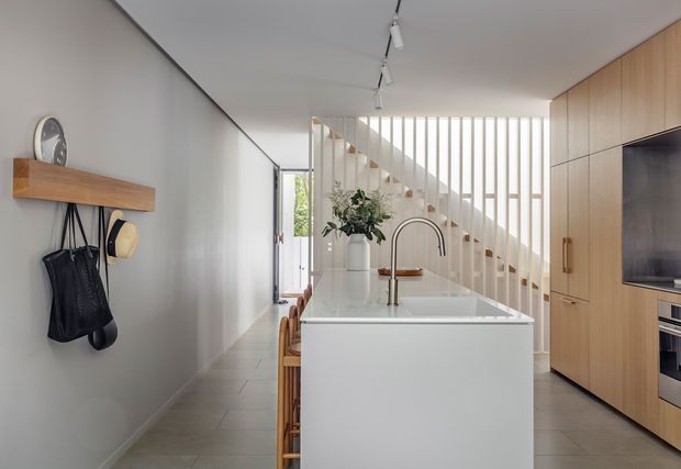 Bright contemporary open-plan kitchen viewed toward a white island, with integrated sink and curved brushed-metal tap, timber bar stools, pale cabinetry with built-in appliances, and a vertical timber stair screen behind. The space has light tiled flooring, white walls and ceiling, track lighting, and a minimalist timber entry shelf with hooks along the left wall.
