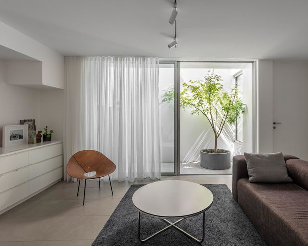 A minimalist contemporary living room with a low-profile brown sectional sofa, round white coffee table, dark area rug, and sculptural timber lounge chair. White sheer curtains cover large sliding glass doors opening to a small internal courtyard with a potted tree. A white built-in cabinet and recessed shelving line the left wall, while track lighting and neutral finishes create a calm, bright interior.