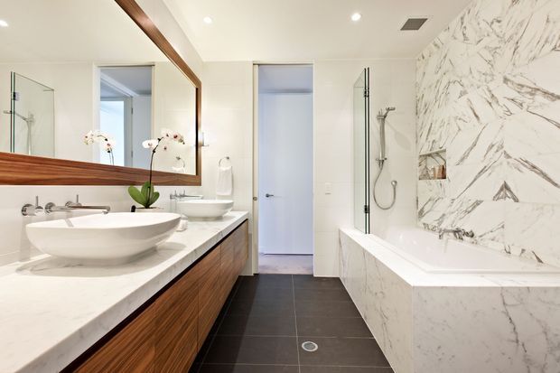 Bright contemporary bathroom featuring a floating dark timber vanity with white vessel basins, a large timber-framed mirror, dark tiled flooring, and a freestanding bathtub set against a dramatic white marble-look feature wall. A glass-enclosed shower and recessed shelving are visible beside the bath.