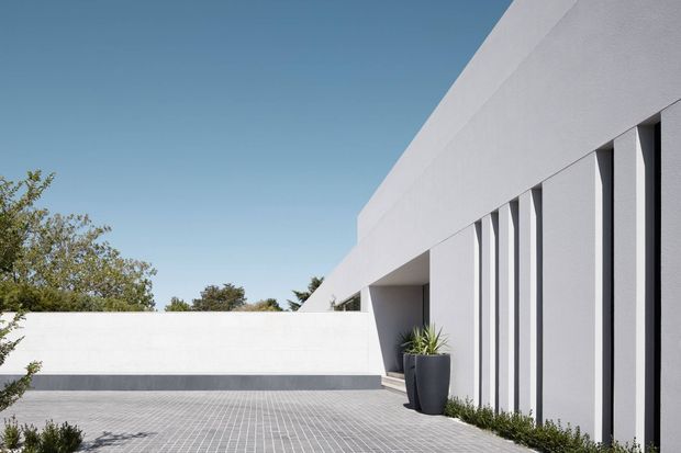 A minimalist modern building exterior with crisp white rendered walls, a long linear facade, deep recessed entry, and closely spaced vertical window or screen elements. The building opens onto a broad grey tiled courtyard or paved forecourt, with restrained landscaping and clear blue sky.