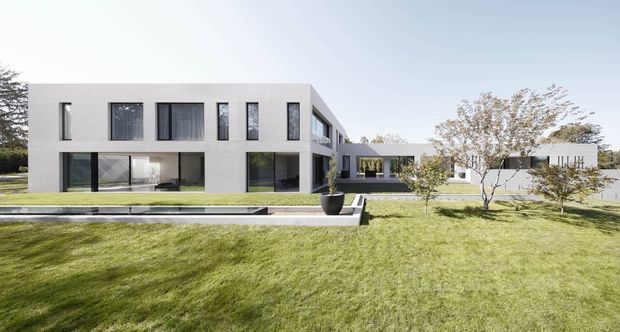 Wide exterior view of a large contemporary two-storey residence with a minimalist rectangular form, pale rendered facade, extensive dark-framed glazing, and a long low connecting wing. The house is set within a spacious landscaped lawn with young trees, clipped grass, a linear reflecting pool or water feature, and a paved terrace.