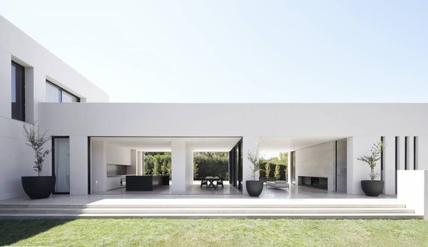 A minimalist modern white rendered house viewed from the lawn, with a long low pavilion-like ground floor opening onto a broad stepped terrace. Full-height glazing and recessed openings reveal a sparse interior with dark kitchen cabinetry and a fireplace, while large black planters with slender trees punctuate the facade. A simple upper-level volume and crisp flat rooflines complete the stark geometric composition.