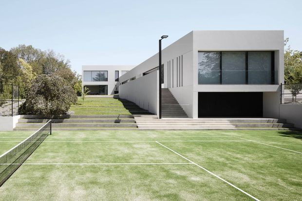 Contemporary white geometric residence viewed from a landscaped tennis court. The low, rectilinear building has a flat roof, broad dark-framed glazing, a recessed garage, external stairs, and a long side wall leading to a second pavilion. Clean lawns, planted borders, and a tennis net occupy the foreground.