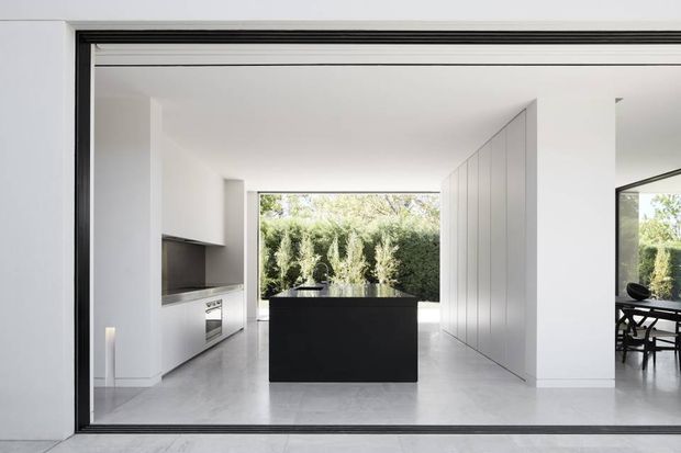 Bright minimalist open-plan kitchen viewed through a large black-framed glazed opening. The room features a central dark island, streamlined white cabinetry, a pale stone or concrete-look floor, integrated appliances, and full-height white wall panelling. A dining area is visible to the right, with a leafy garden beyond the rear glazing.