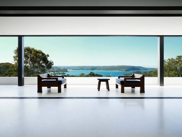 A minimalist living room with expansive floor-to-ceiling glazing framing a bright coastal water view. Two dark timber lounge chairs with light cushions face each other across a small dark wood side table. The room has a white interior, polished pale floor, strong horizontal lines, and dark slender window frames and structural columns.