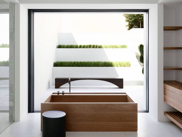A bright, minimalist bathroom featuring a freestanding rectangular timber bathtub positioned in front of a large black-framed window. A wall-mounted black faucet and simple dark bath screen detail sit above the tub. Built-in timber shelving and drawers are visible on the right, while a black cylindrical stool provides contrast. Through the window is a clean white exterior courtyard with tiered planted ledges.