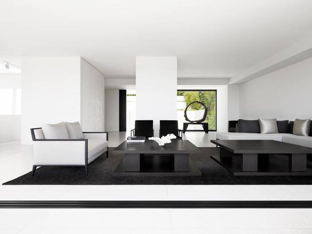 A bright, minimalist open-plan living and dining space with a monochrome palette. White walls, ceiling and large-format pale flooring frame a black dining table, black-framed armchair, dark low coffee tables and a dark rug. A light upholstered sofa and armchair provide contrast, while floor-to-ceiling glazing at the rear opens to greenery and a sculptural circular object. Clean-lined built-in joinery and sparse furnishings create a refined contemporary interior.