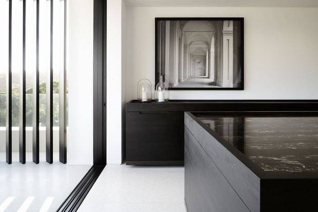 A refined contemporary interior featuring a dark timber or black-stained built-in sideboard and a large island or counter in the foreground. White walls and a pale glossy floor create a bright, high-contrast setting, while full-height glazing with slim black vertical frames opens to greenery. A monochrome architectural photograph and minimal decorative objects add a restrained, gallery-like character.