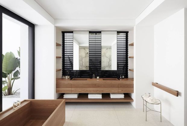 Bright contemporary bathroom featuring a freestanding timber bathtub in the foreground and a floating timber vanity with integrated basins, large mirrors, and black horizontal window screens behind it. White walls, a pale tiled floor, timber shelving, and black-framed glazing create a clean, refined finish.